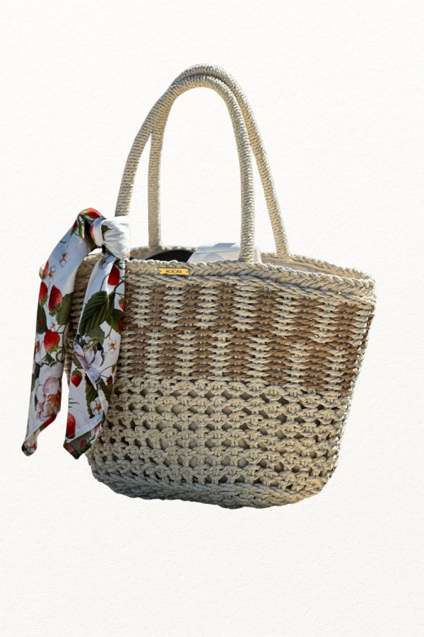 Cesto de palha à beira-mar com lenço de morangos removível, segurado por uma mão com pulseira delicada.