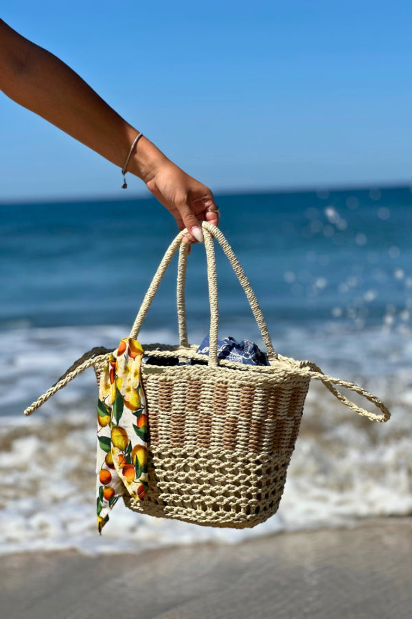 Cesto de palha à beira-mar com lenço de limões removível, segurado por uma mão com pulseira delicada.