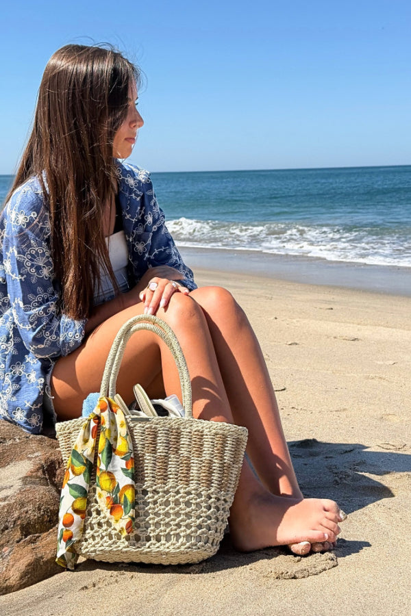 Cesto de palha à beira-mar com lenço de limões removível, segurado por uma mão com pulseira delicada.