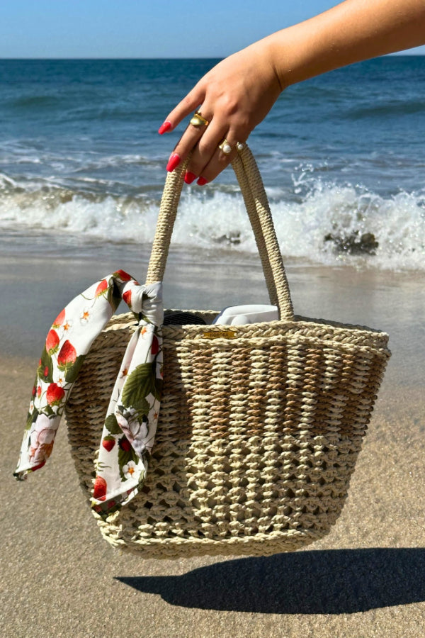 Cesto de palha à beira-mar com lenço de morangos removível, segurado por uma mão com pulseira delicada.