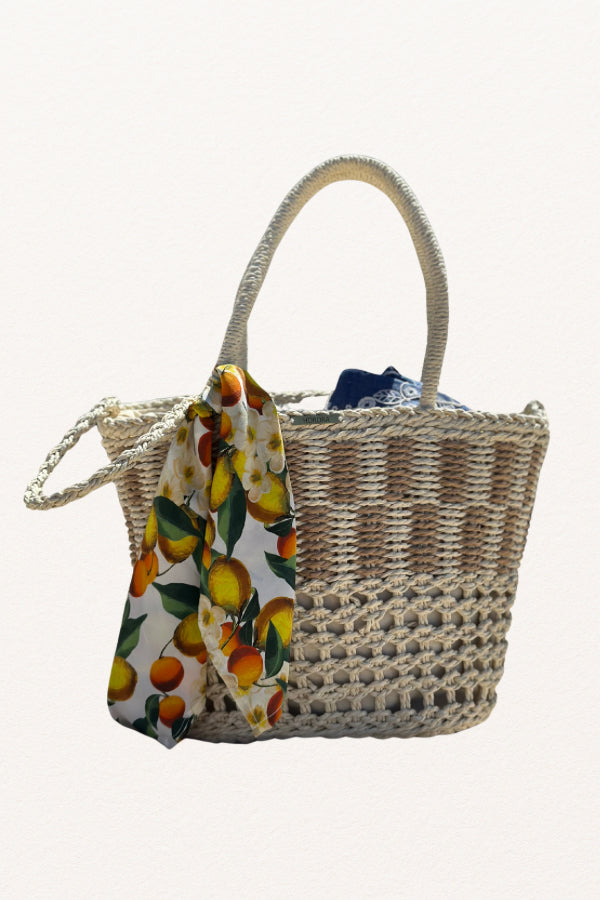Cesto de palha à beira-mar com lenço de limões removível, segurado por uma mão com pulseira delicada.