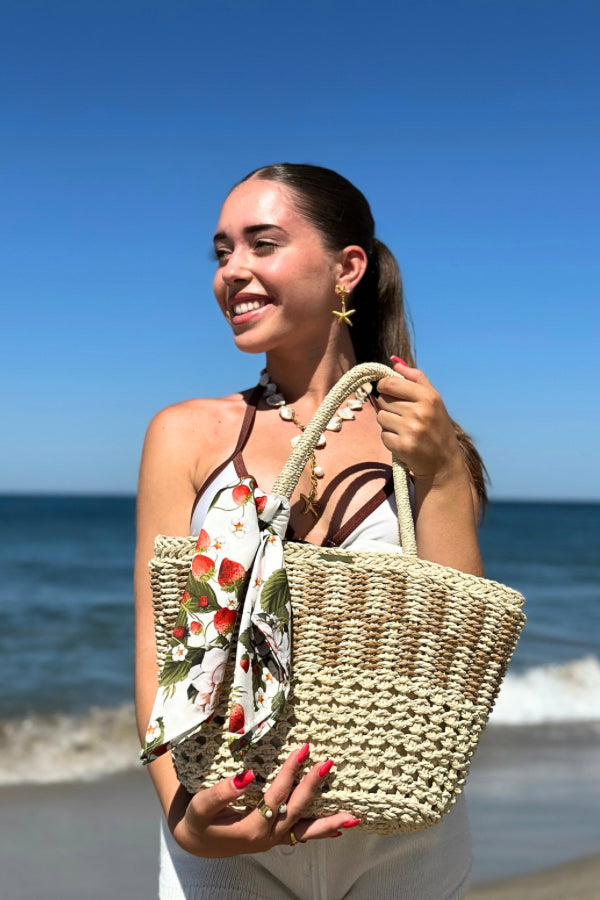 Cesto de palha à beira-mar com lenço de limões removível, segurado por uma mão com pulseira delicada.