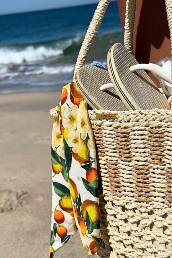 Cesto de palha à beira-mar com lenço de limões removível, segurado por uma mão com pulseira delicada.
