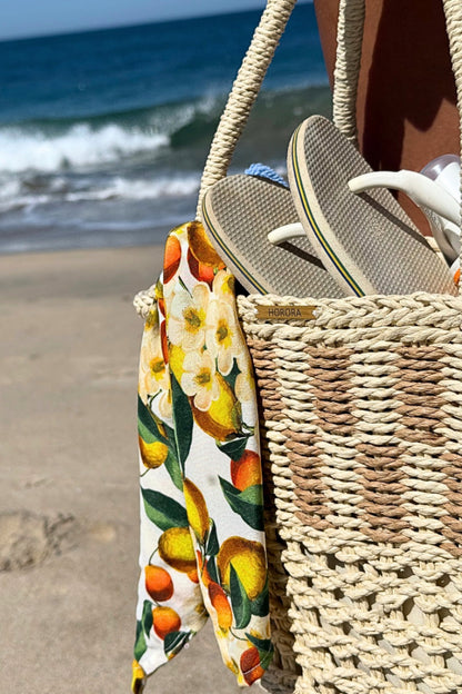 Cesto de palha à beira-mar com lenço de limões removível, segurado por uma mão com pulseira delicada.